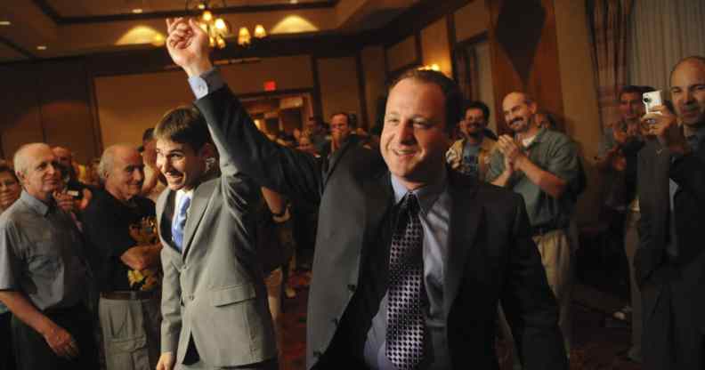 Jared Polis (R) and Marlon Reis hold hands as people clap behind them