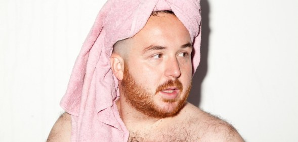 Scottee, shirtless, wearing a pink towel on his head