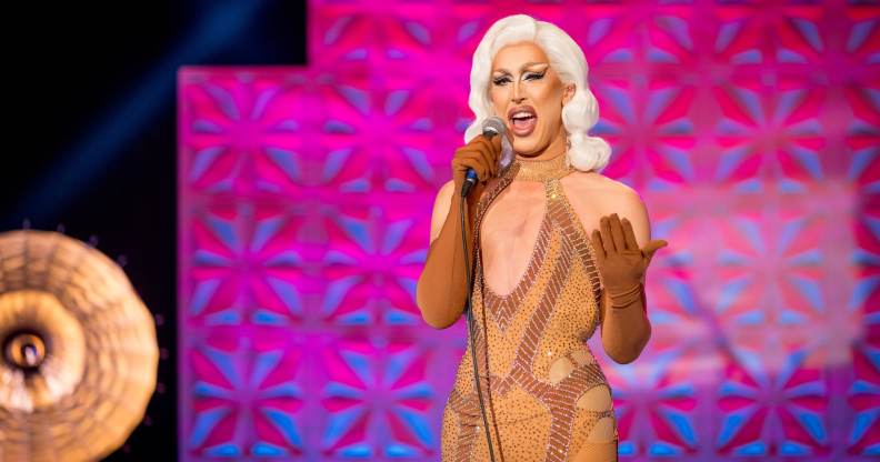 A'Whora, a drag queen in a flesh-coloured, stoned body con dress and blonde hair, performing stand up into a mic