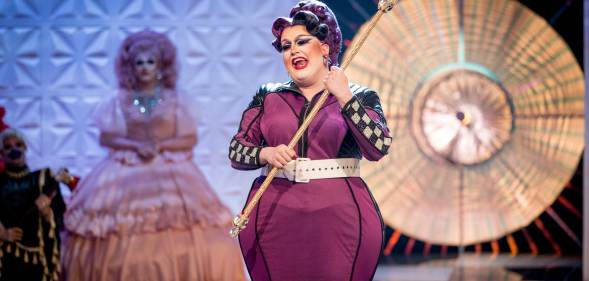 Lawrence Chaney, a drag queen in a long purpler dress with racing flag tripe down the sides, holding a sceptre