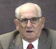 Dick Marston in a grey suit and red tie speaks to the camera against a grey background