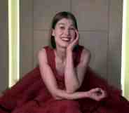 Rosamund Pike sitting in a giant red tule dress, resting her head on her hand and smiling