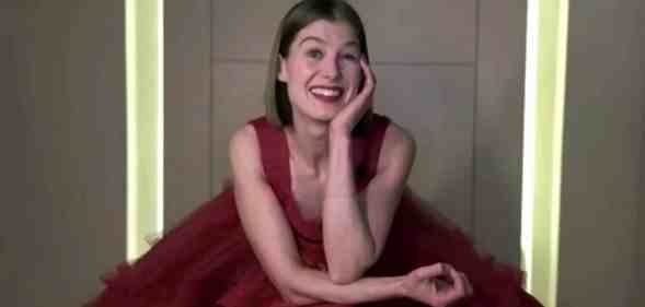 Rosamund Pike sitting in a giant red tule dress, resting her head on her hand and smiling
