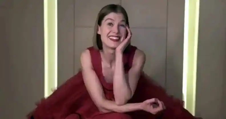 Rosamund Pike sitting in a giant red tule dress, resting her head on her hand and smiling