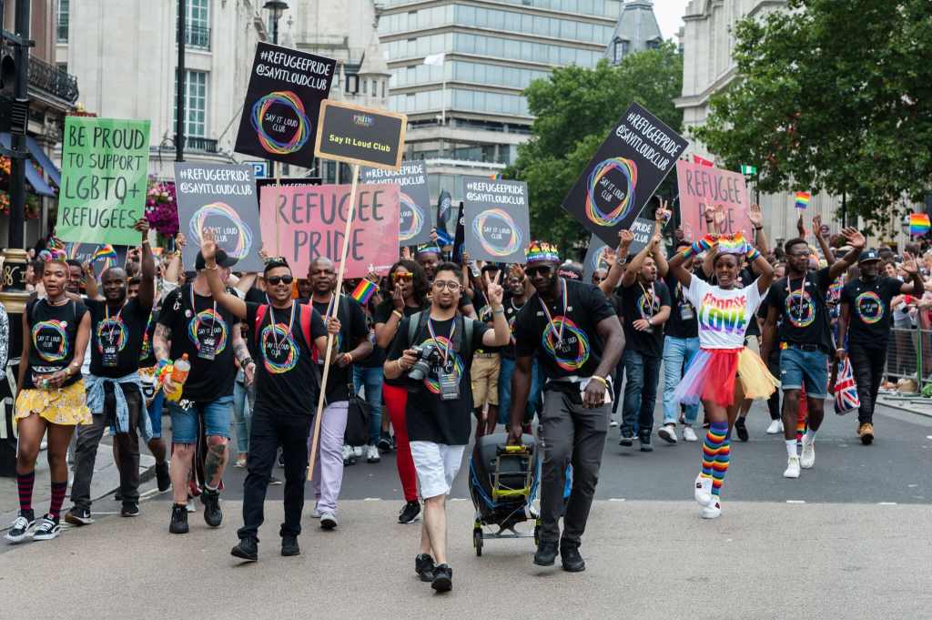 A crowd of mostly Black and brown LGBT+ protesters holding placards with messages like "refugee pride" and "be proud to support LGBTQ refugees"