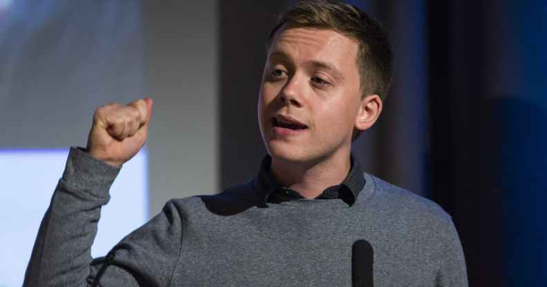 Owen Jones speaks in front of a podium in a grey jumper over a blue shirt