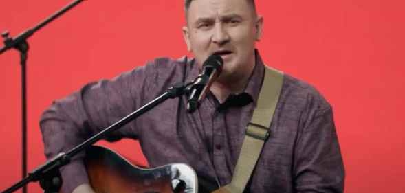 Dmitry Butakov sings while strumming the guitar against a red background