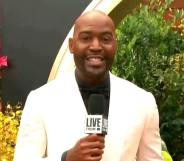 Karamo Brown holds a microphone in a white blazer and black shirt on the Oscars red carpet