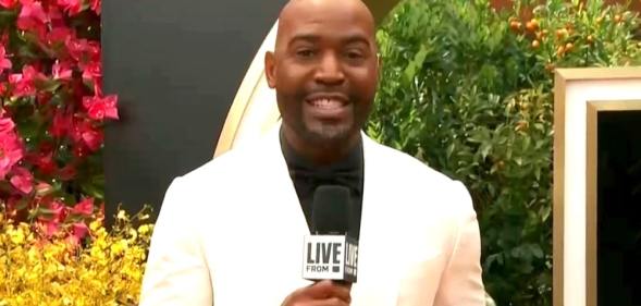 Karamo Brown holds a microphone in a white blazer and black shirt on the Oscars red carpet