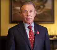 Charles Peruto stands in front of a painting in a red tie and dark suit