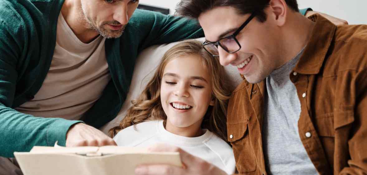 Happy gay parents reading book together with their little daughter at home
