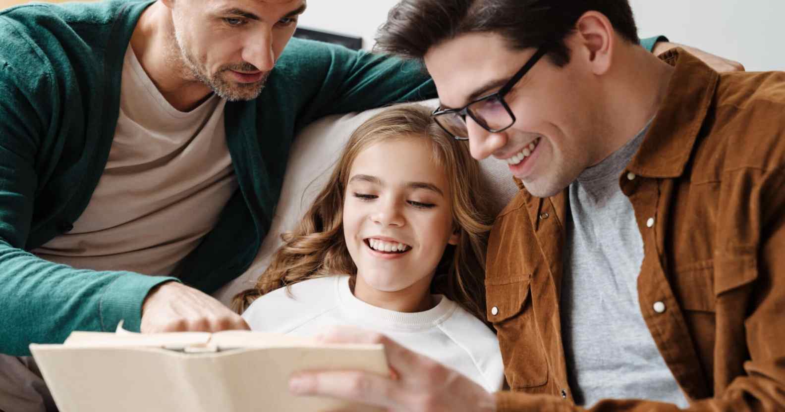 Happy gay parents reading a book together with their little daughter at home