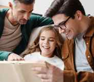 Happy gay parents reading book together with their little daughter at home