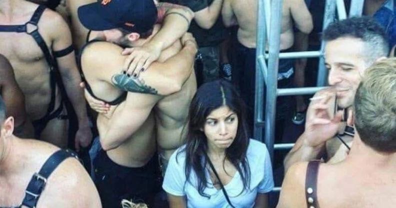 A young woman in a v-neck t-short surrounded by leather-clad, shirtless men