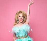 Miz Cracker in a fluffy blue gown against a pink background, with one hand in the air, smiling