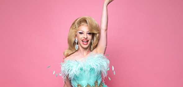 Miz Cracker in a fluffy blue gown against a pink background, with one hand in the air, smiling