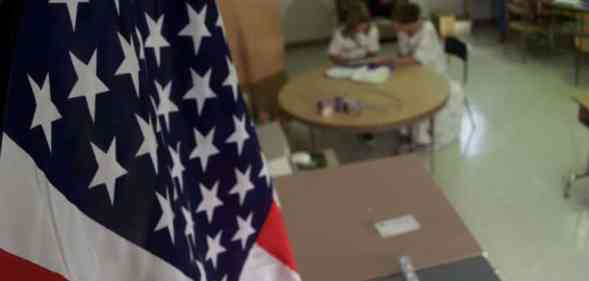 American flag hangs in the foreground of a classroom as two people sit at a table in the background