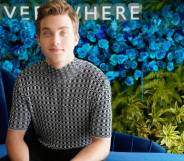 Actor Jordan Elsass sits on a blue couch in front of a blue background with green plants