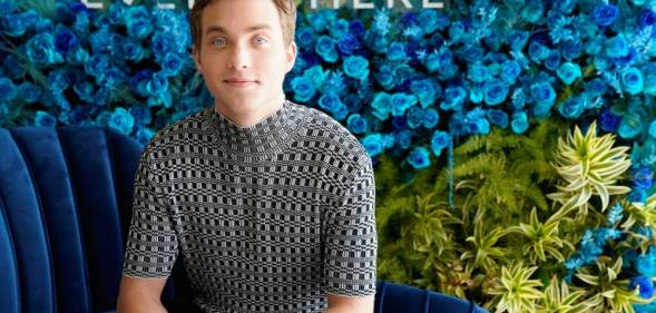 Actor Jordan Elsass sits on a blue couch in front of a blue background with green plants