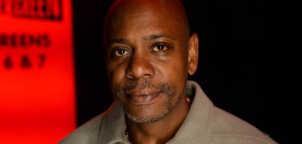 Dave Chappelle wears a neutral coloured shirt amid a black and red background