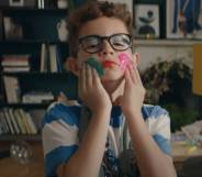 still from the latest John Lewis home insurance ad. The still depicts a young boy wearing a dress and colourful makeup smearing paint on his face
