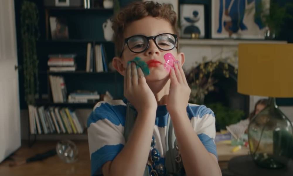 still from the latest John Lewis home insurance ad. The still depicts a young boy wearing a dress and colourful makeup smearing paint on his face
