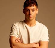 British Olympian Tom Daley stands in front of a neutral coloured background with his arms crossed