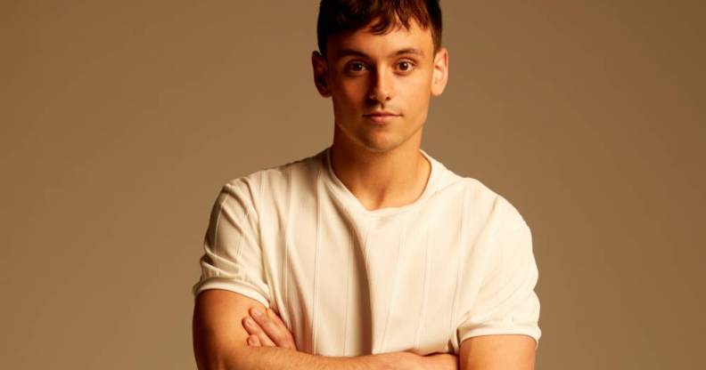 British Olympian Tom Daley stands in front of a neutral coloured background with his arms crossed