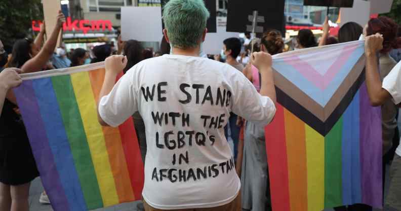 Person stood with their back to the camera. The back of their top reads: "We stand with LGBTQs in Afghanistan". They're holding a rainbow Pride flag and the Progress Pride flag