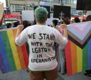 Person stood with their back to the camera. The back of their top reads: "We stand with LGBTQs in Afghanistan". They're holding a rainbow Pride flag and the Progress Pride flag