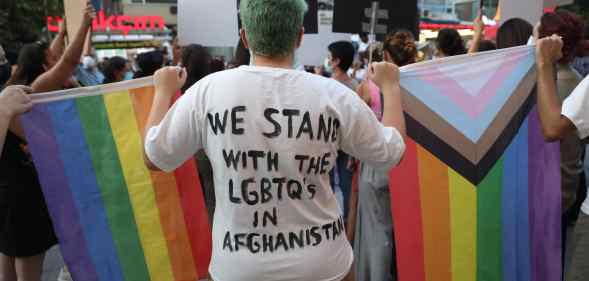 Person stood with their back to the camera. The back of their top reads: "We stand with LGBTQs in Afghanistan". They're holding a rainbow Pride flag and the Progress Pride flag