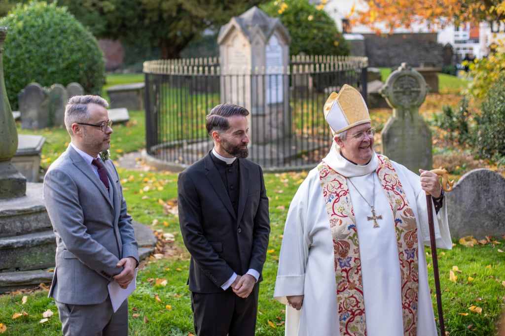 Male in grey suit and male in vicar's attire receive blessing from bishop