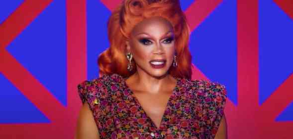 RuPaul appears in a red wig and red and blue outfit in front of a geometric patterned background