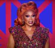 RuPaul appears in a red wig and red and blue outfit in front of a geometric patterned background