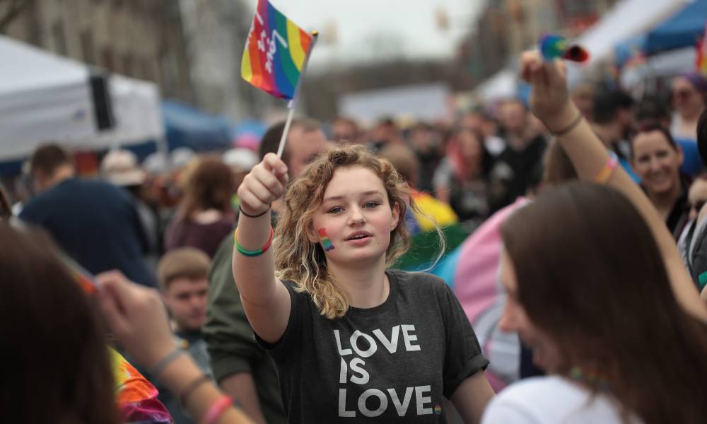 Young person holds up an LGBT+ pride flag while wearing a shirt that says 'love is love'