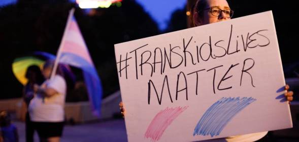 An image shows a trans rights advocate holding a sign that reads 'trans kids lives matter'