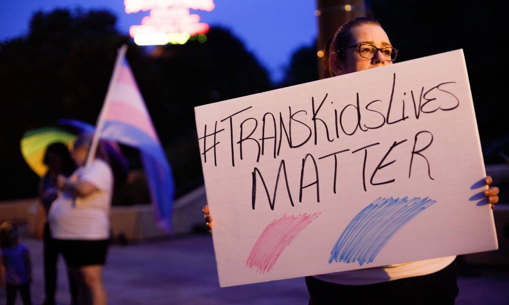 An image shows a trans rights advocate holding a sign that reads 'trans kids lives matter'