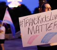 An image shows a trans rights advocate holding a sign that reads 'trans kids lives matter'