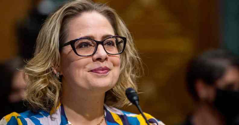 Kyrsten Sinema wears a black, blue, yellow and white patterned top while speaking into a microphone at a US Senate Committee on Finance hearing