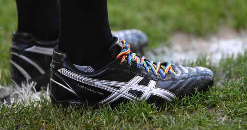 A person wearing black trainers stands on a football pitch. The trainers have rainbow laces to show support for the LGBT+ community