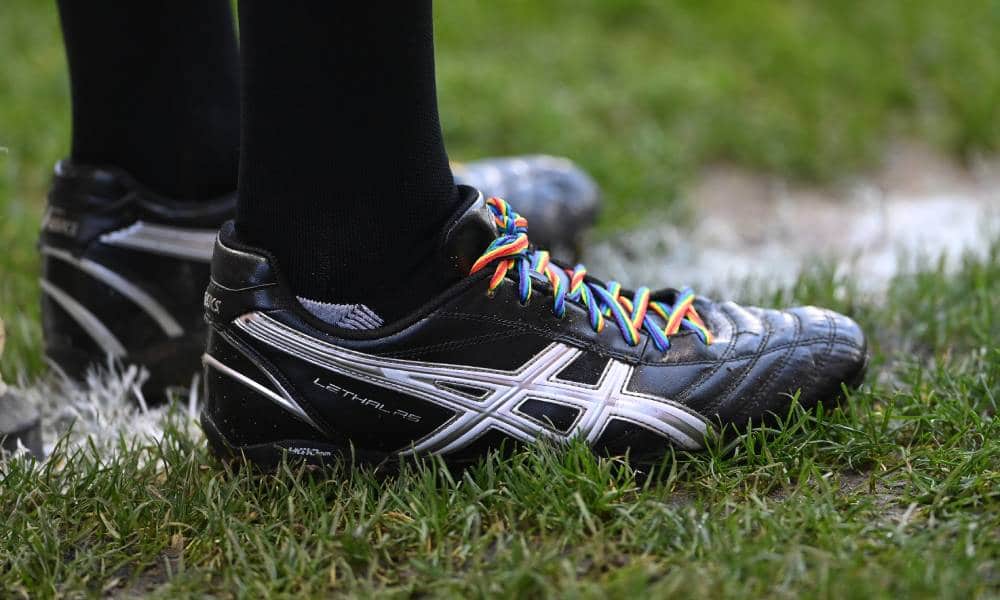 A person wearing black trainers stands on a football pitch. The trainers have rainbow laces to show support for the LGBT+ community
