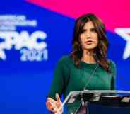 Kristi Noem wears a green outfit and stands at a podium in front of a red and blue background