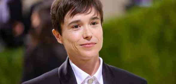 Elliot Page smiles at the camera while wearing a white button up shirt and black suite jacket