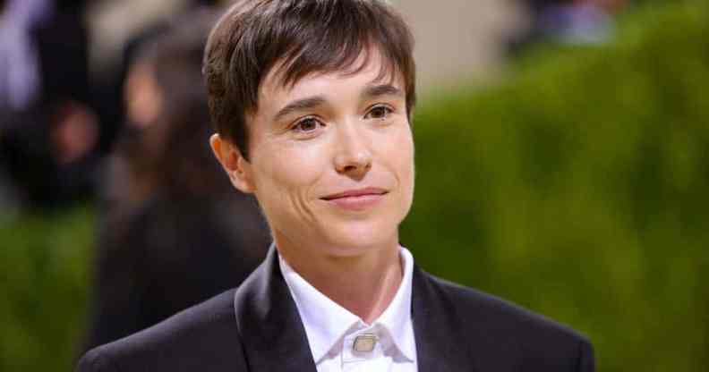 Elliot Page smiles at the camera while wearing a white button up shirt and black suite jacket