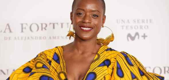 T'Nia Miller smiles at the camera while wearing a beautiful yellow, brown and blue dress and gold hoop earrings