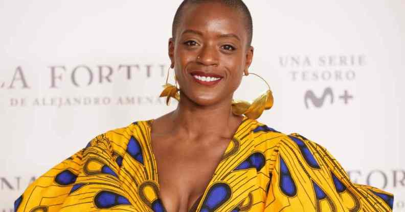 T'Nia Miller smiles at the camera while wearing a beautiful yellow, brown and blue dress and gold hoop earrings
