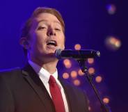 Clay Aiken sings into a microphone as he wears a white shirt, black jacket and red tie
