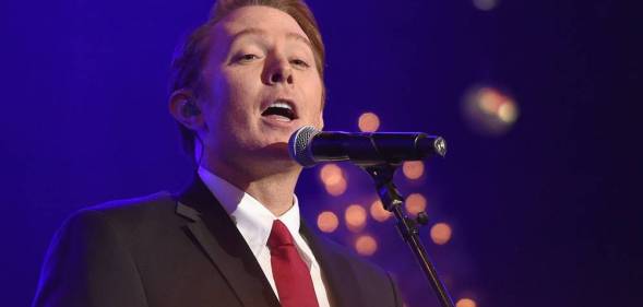 Clay Aiken sings into a microphone as he wears a white shirt, black jacket and red tie