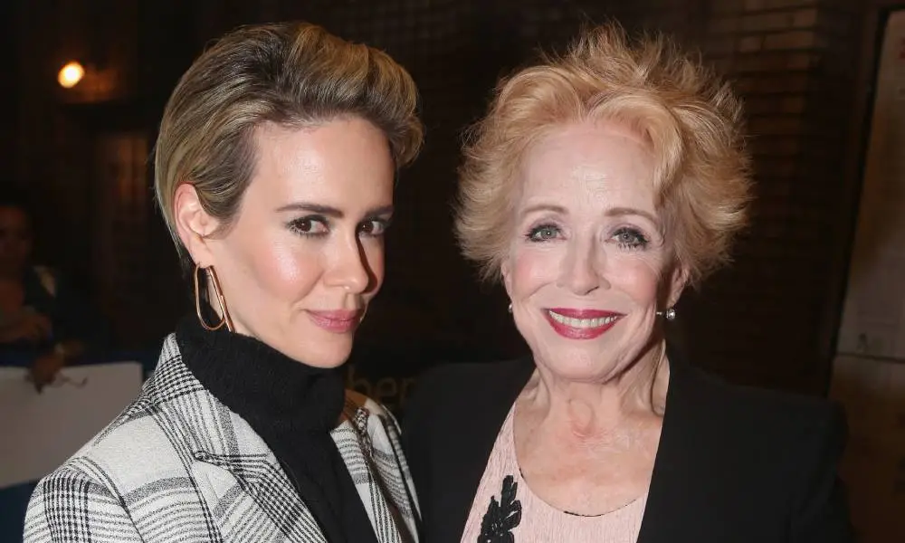 Sarah Paulson and Holland Taylor pose for the camera. Paulson is wearing a black turtle neck, grey plaid jacket and hoop earrings. Taylor is wearing a nude coloured shirt with a black jacket