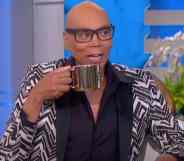 RuPaul appears on the Ellen DeGeneres show in a black, white and grey patterned suit, holding a gold coloured mug